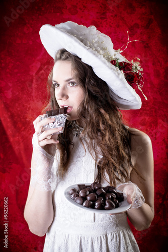 Beautiful young lady in retro clothes eating chocolate on red copy space background and looking forward