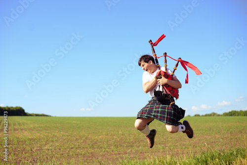 Picture of male jumping high with pipes in Scottish traditional kilt on green outdoors copy space summer field background