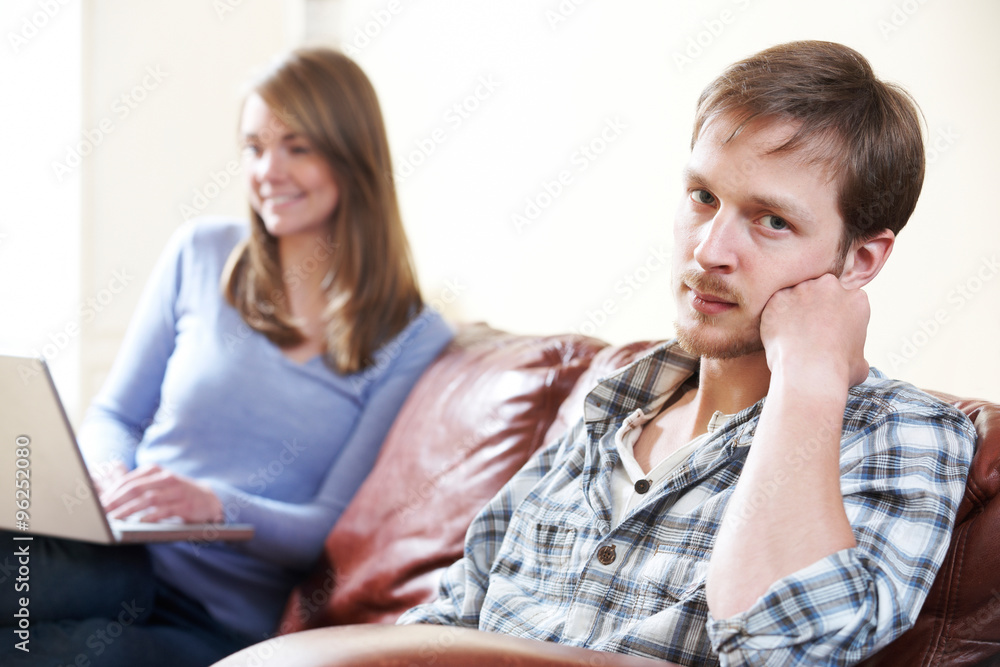 Unhappy Man Sitting On Sofa As Partner Uses Computer Unhappy Man Sitting On Sofa As Partner Uses Computer