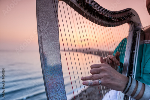 Fototapet Close up of the hands of woman playing harp by the sea at sunset