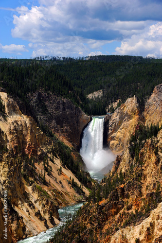 Big Spray from Upper Falls in Yellowstone National Park shows up against the sheer sandstone cliffs of Yellowstone Canyon. Blue skies are overhead.