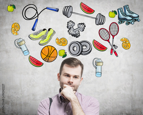 A portrait of a dreaming handsome man who is thinking about his choice of sport activity. Colourful sport icons are drawn on the concrete wall. A concept of a healthy lifestyle.