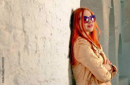 Redhair woman looking away happy. Trendy girl posing near white brick wall in casual clothes and fashionable 60th style sunglasses. A lot of space for text. Instagram color imitation.