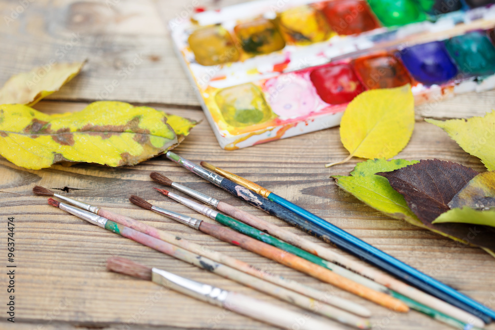 Plastic paint palette with paint, brushes and yellow leaves on w