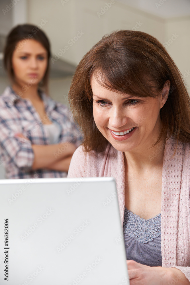 Mother Using Laptop With Frustrated Teenage Daughter In Backgrou Mother Using Laptop With Frustrated Teenage Daughter In Backgrou