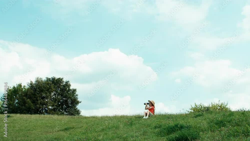 Young naturalist through binoculars watching wildlife and looking at camera. Girl sitting on the grass in the hill against the sky and waving hand at camera Young naturalist through binoculars watching wildlife and looking at camera. Girl sitting on the grass in the hill against the sky and waving hand at camera