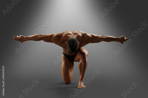 muscular male model stands on one knee, arms outstretched to the side and looks down