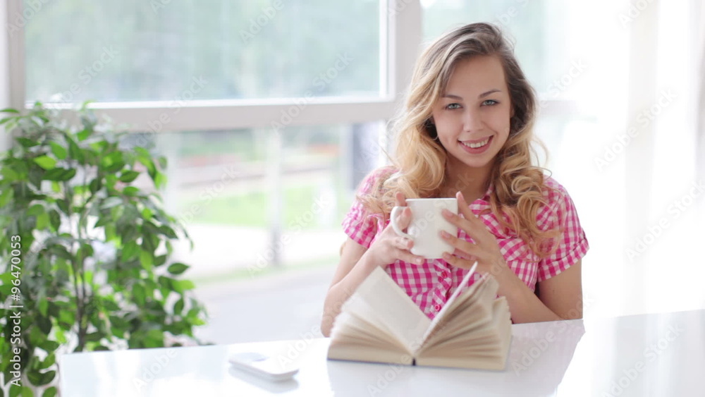 Pretty blond girl sitting at table with opened book smiling and drinking from cup Pretty blond girl sitting at table with opened book smiling and drinking from cup
