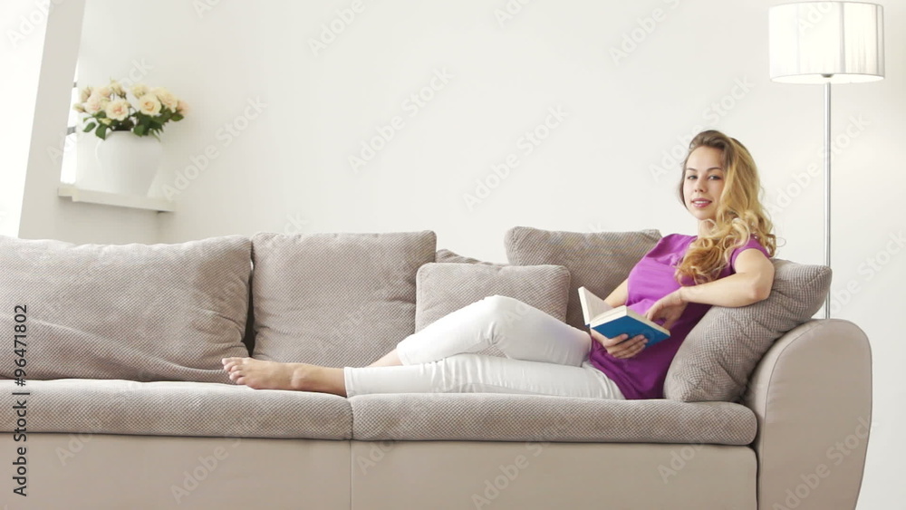 Pretty young woman lying on sofa reading book and looking at camera with smile Pretty young woman lying on sofa reading book and looking at camera with smile
