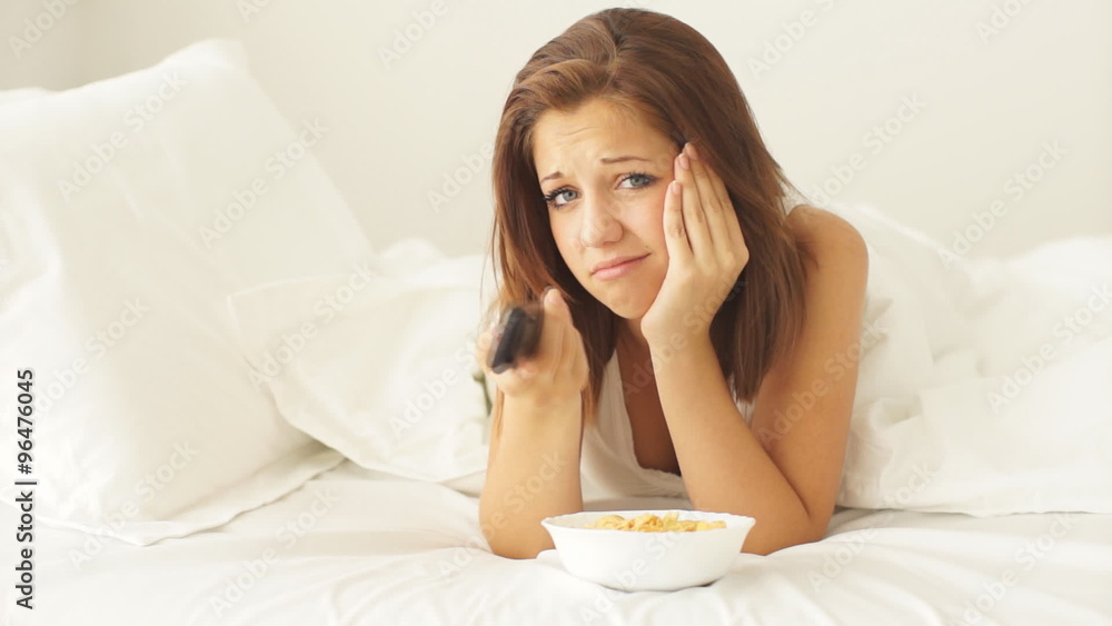 Pretty girl lying in bed pressing buttons on remote control eating corn flakes and smiling at camera Pretty girl lying in bed pressing buttons on remote control eating corn flakes and smiling at camera