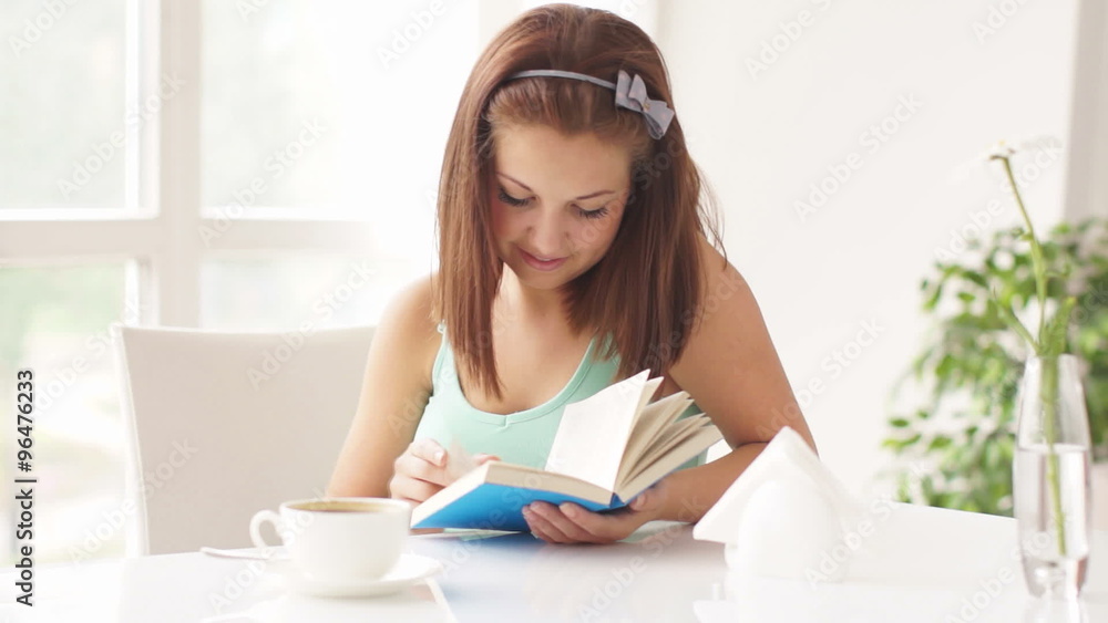 Pretty smiling girl sitting at table reading book and looking at camera Pretty smiling girl sitting at table reading book and looking at camera