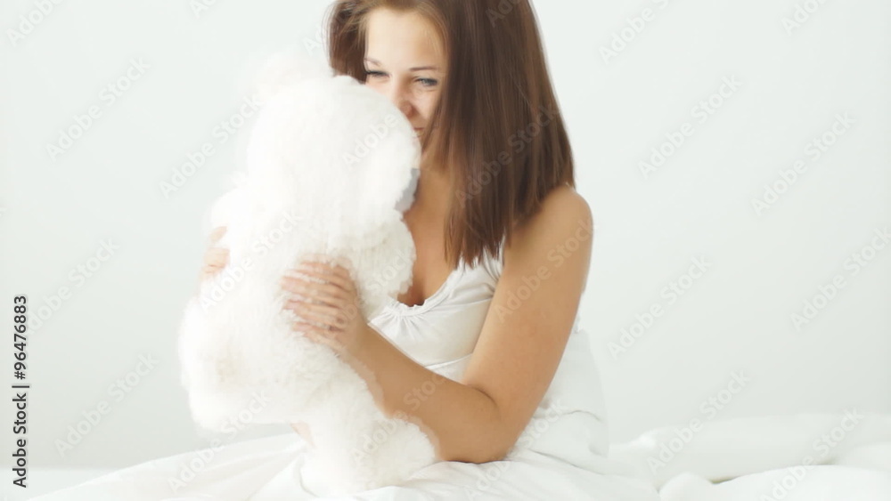 Young woman sitting on bed playing and cuddling with teddy bear and smiling at camera Young woman sitting on bed playing and cuddling with teddy bear and smiling at camera