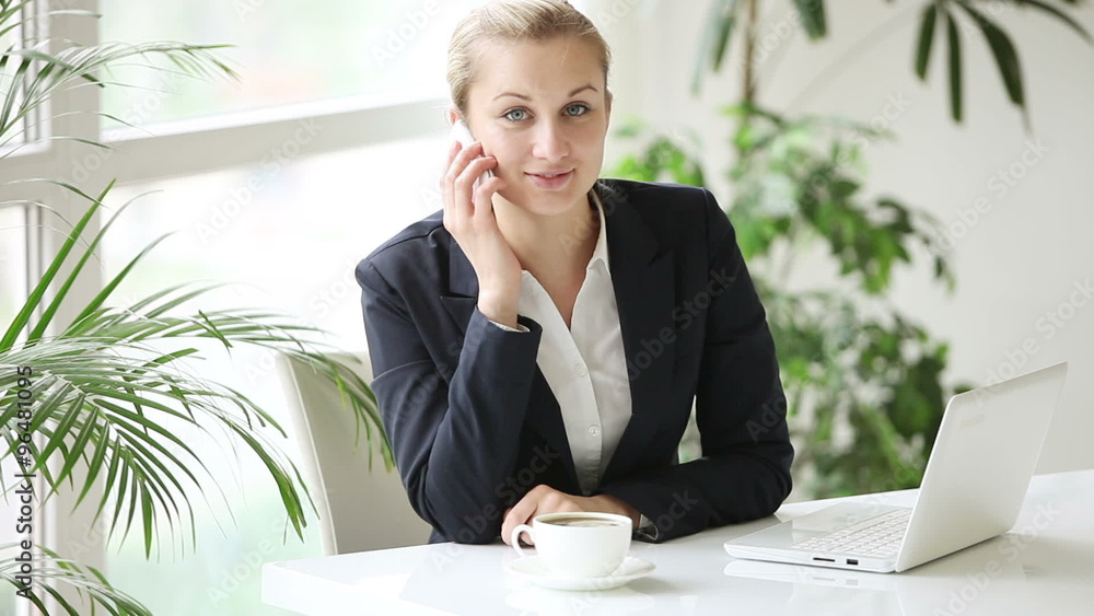 Young businesswoman sitting at office table talking on mobile phone and looking at camera with smile Young businesswoman sitting at office table talking on mobile phone and looking at camera with smile