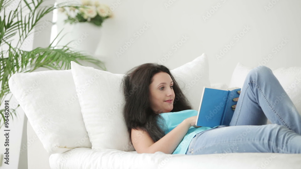Attractive young woman lying on sofa reading book and smiling at camera Attractive young woman lying on sofa reading book and smiling at camera