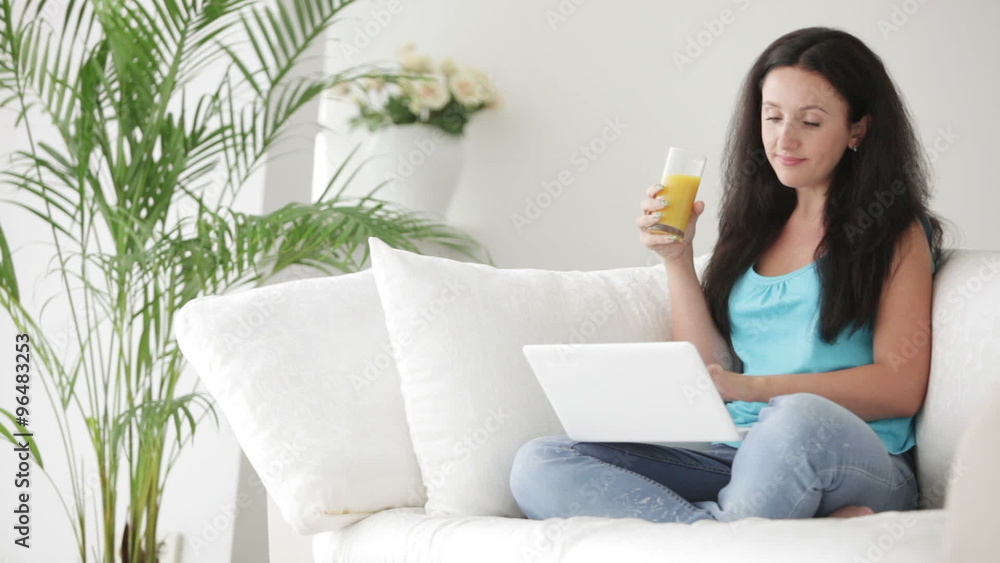 Pretty brunette woman sitting on sofa using laptop drinking juice and smiling at camera Pretty brunette woman sitting on sofa using laptop drinking juice and smiling at camera