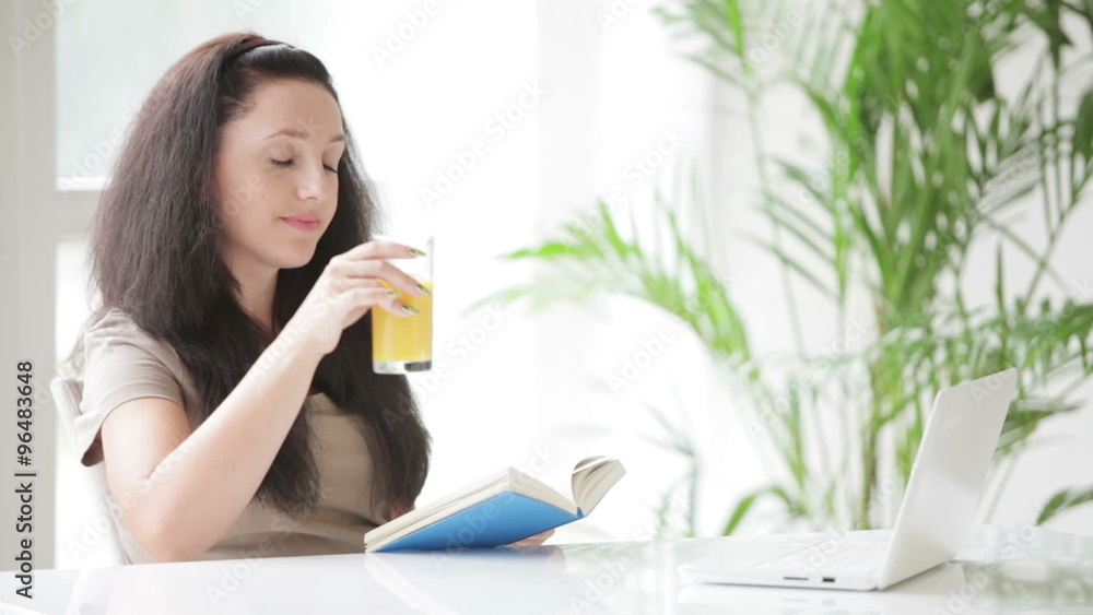 Smiling young woman sitting at table drinking juice reading book and looking at camera Smiling young woman sitting at table drinking juice reading book and looking at camera