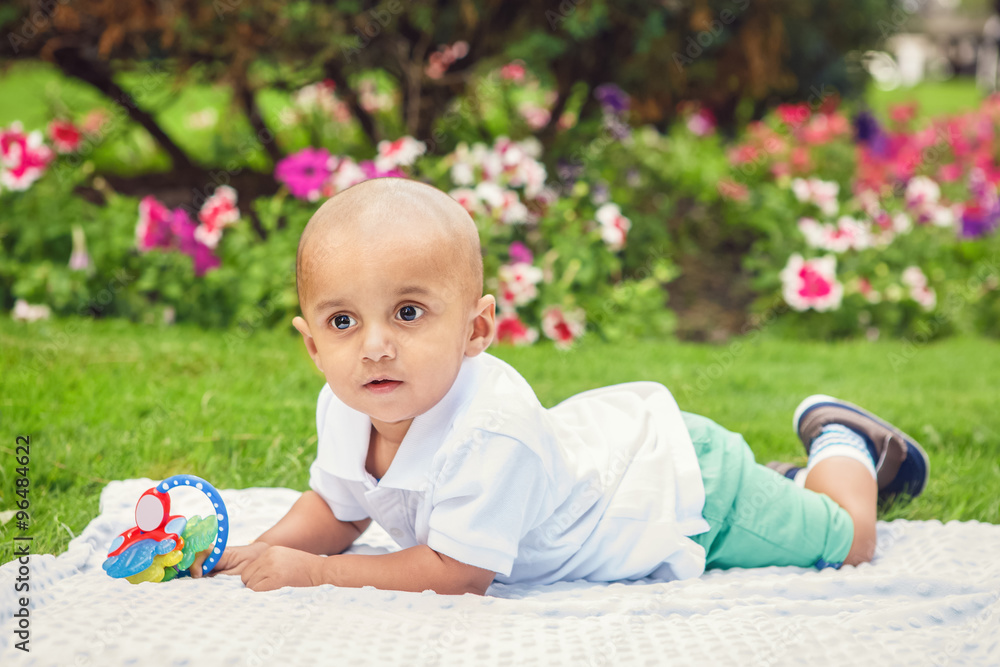 Portrait of cute adorable little indian South Asian or Middle Eastern infant boy in white shirt laying on ground in park outside on bright summer day smiling and playing Portrait of cute adorable little indian South Asian or Middle Eastern infant boy in white shirt laying on ground in park outside on bright summer day smiling and playing