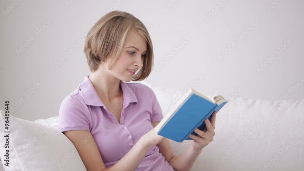 Pretty young woman sitting on sofa reading book looking at camera and smiling Pretty young woman sitting on sofa reading book looking at camera and smiling