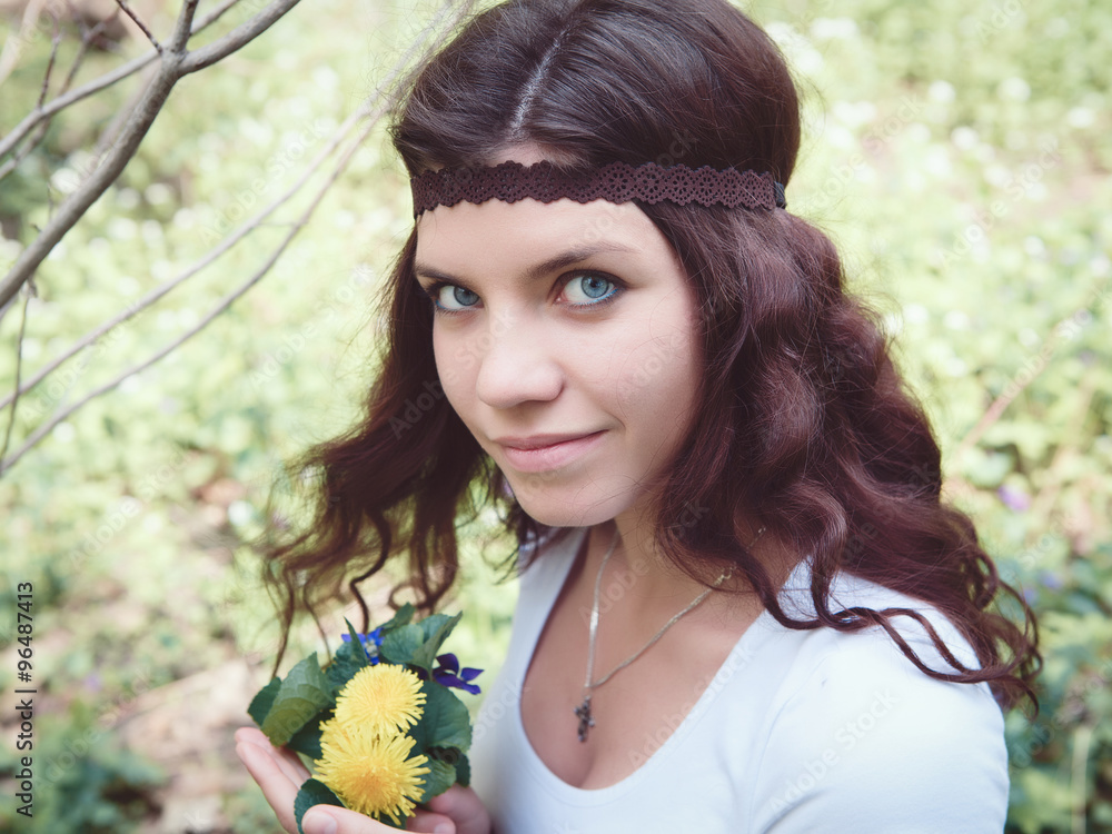 Naklejka premium Portrait of beautiful Caucasian girl with long red hair in white top with brown hippie style headband, with yellow flowers dandelions in forest park outside, summer spring day looking in camera