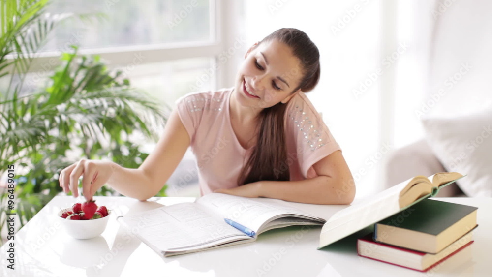 Cute girl studying at table with books and eating strawberries Cute girl studying at table with books and eating strawberries