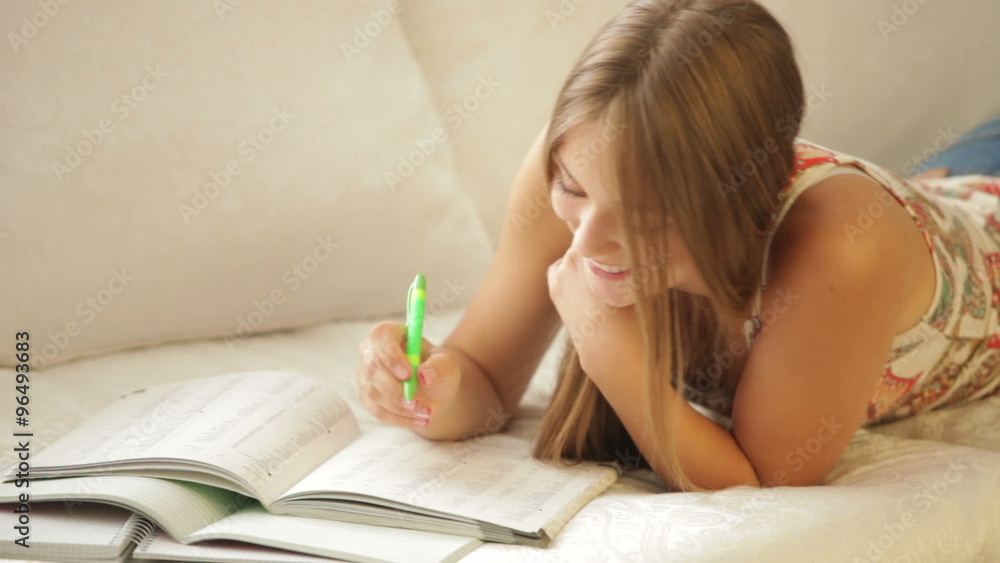 Cheerful girl lying on sofa writing in notebook and smiling Cheerful girl lying on sofa writing in notebook and smiling