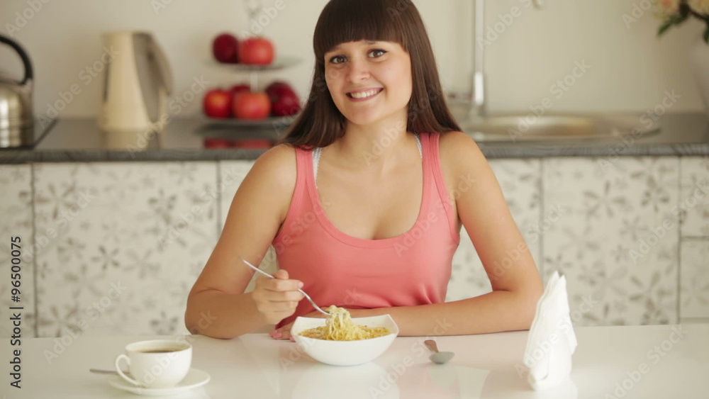Cheerful girl sitting at kitchen table and eating noodle Cheerful girl sitting at kitchen table and eating noodle