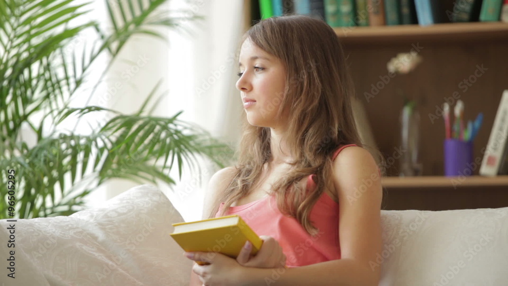 Cheerful girl relaxing on couch holding book looking at camera and smiling. Panning camera Cheerful girl relaxing on couch holding book looking at camera and smiling. Panning camera