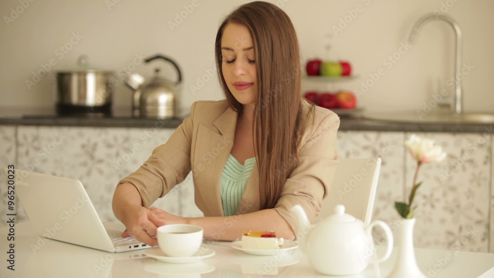 Attractive young woman sitting at table working on laptop drinking tea and smiling at camera Attractive young woman sitting at table working on laptop drinking tea and smiling at camera