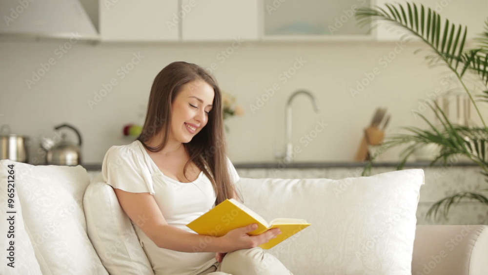 Beautiful girl sitting on sofa reading book looking at camera and smiling