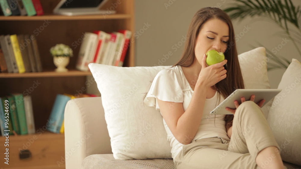 Pretty girl sitting on sofa using touchpad eating apple and smiling