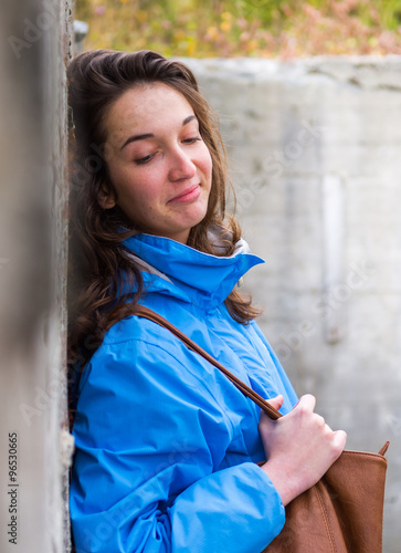 Frau mit braunen haaren und Handtasche lehnt sich gegen eine Betonwand, lächelt mit geschlossenen Augen, vertical