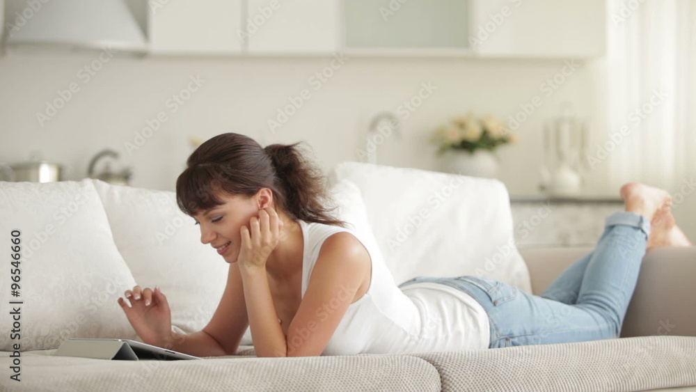 Young woman lying on couch using touchpad and smiling Young woman lying on couch using touchpad and smiling