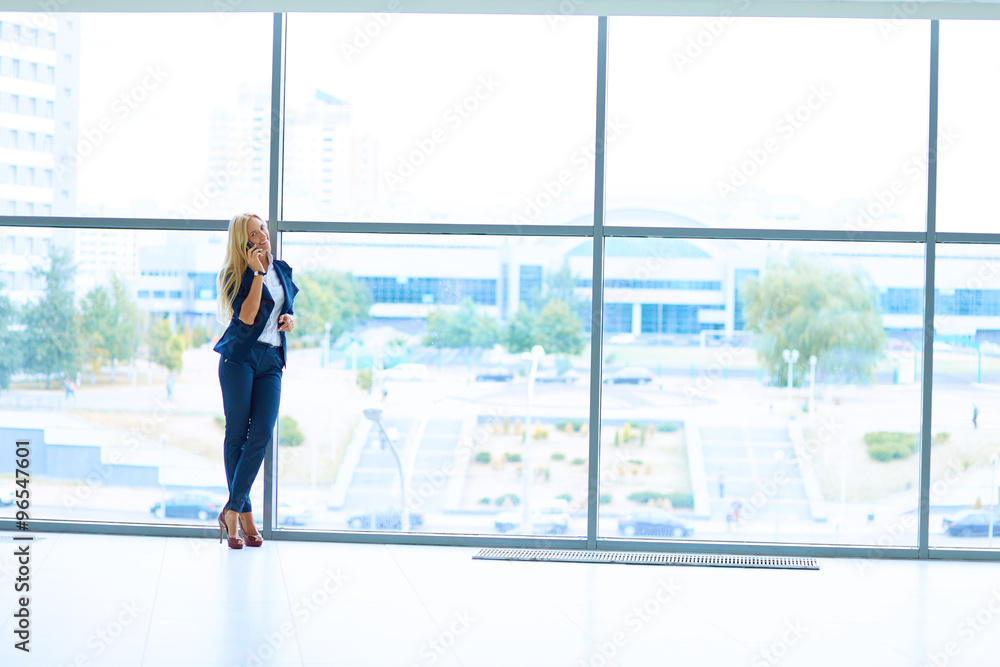 Businesswoman standing against office window talking on mobile phone Businesswoman standing against office window talking on mobile phone