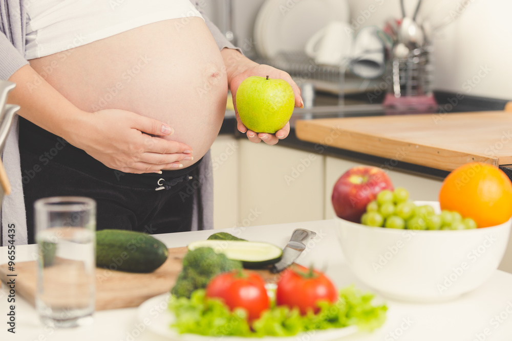 Toned shot of pregnant woman posing with green apple while cooki