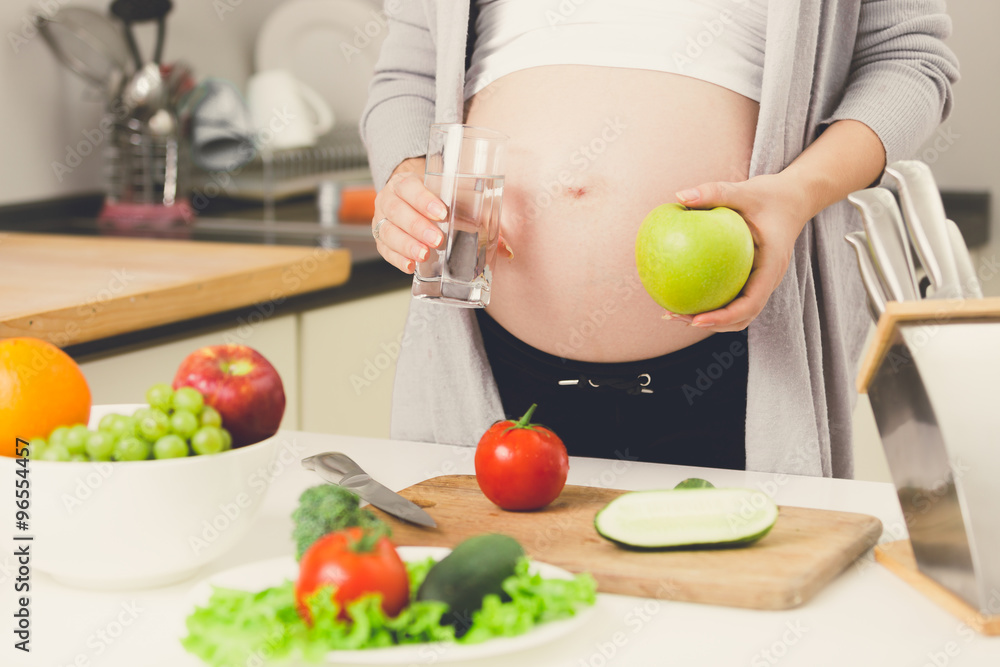 Closeup of pregnant woman posing on kitchen with apple and glass