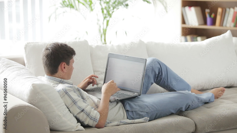 Young man lying on sofa working on laptop looking at camera and smiling Young man lying on sofa working on laptop looking at camera and smiling
