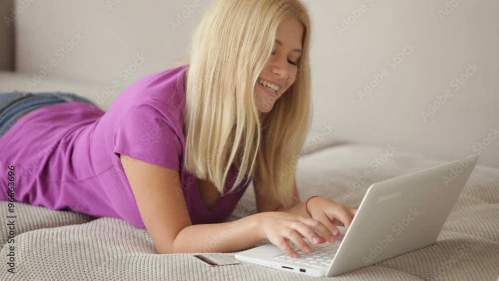 Cheerful girl lying on sofa using laptop looking at camera and smiling. Panning camera Cheerful girl lying on sofa using laptop looking at camera and smiling. Panning camera