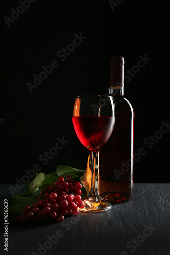 A bottle and a glass of wine, red grapes, on grey-black background