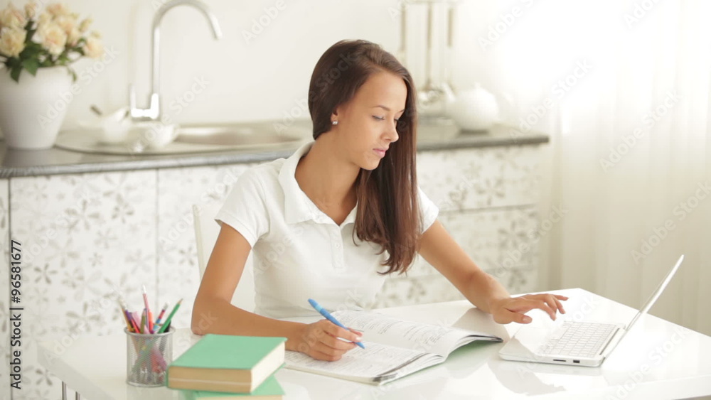 Pretty girl sitting at table writing in workbook using laptop looking at camera and smiling Pretty girl sitting at table writing in workbook using laptop looking at camera and smiling