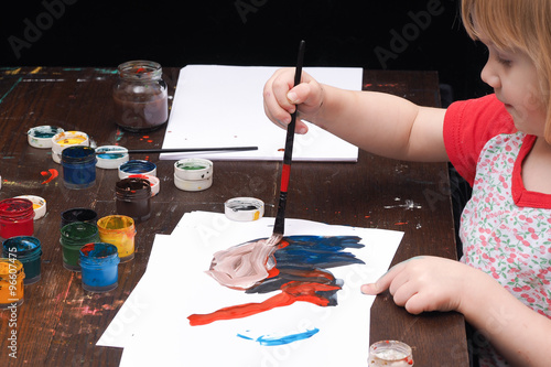 Маленький ребенок рисует красками, гуашью. Темный стол в краске, банки с краской.  Девочка рисует кистью на белой бумаге. Черный фон. У девочки светлые волосы, она очень мала 