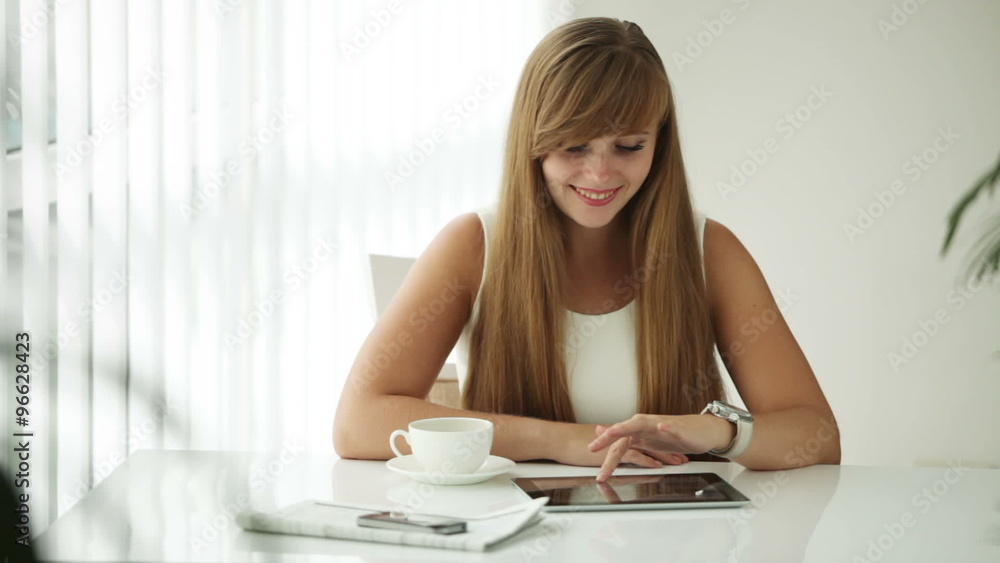Beautiful girl sitting at table drinking coffee using touchpad looking at camera and smiling Beautiful girl sitting at table drinking coffee using touchpad looking at camera and smiling