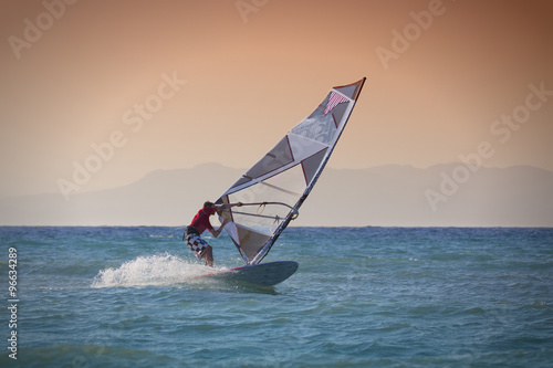Windsurfen in Rhodos Griechenland. Mann surft im blauen Meer vor rötlichem Himmel, Insel im Hintergrund.
