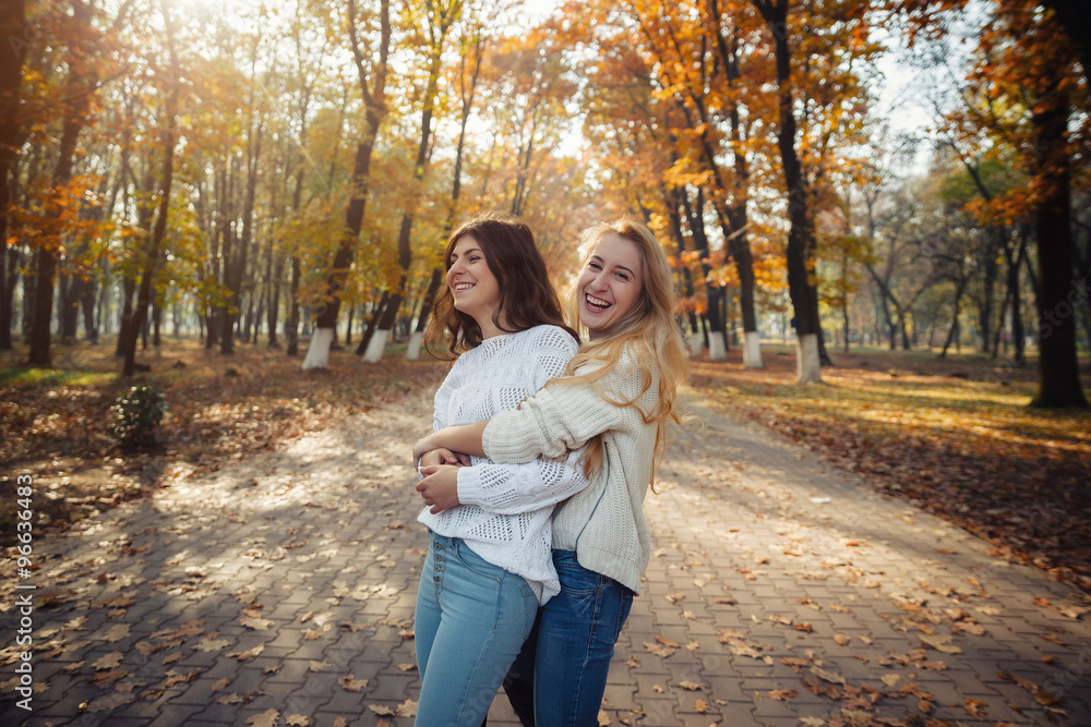 Fototapeta premium Two happy and cheerful young girl students, blond and brunette in coat and jeans laughing in sunny autumn park full of fallen leaves, horizontal pictures
