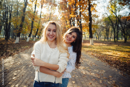 Two happy and cheerful young girl students, blond and brunette in coat and jeans laughing in sunny autumn park full of fallen leaves, horizontal pictures