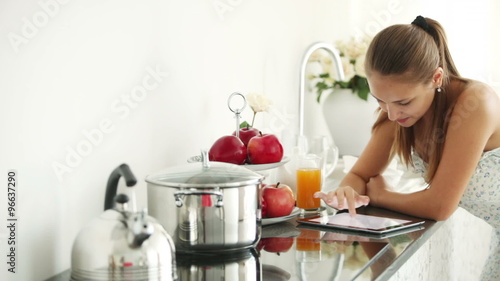 Cheerful girl standing in kitchen using touchpad looking at camera and smiling