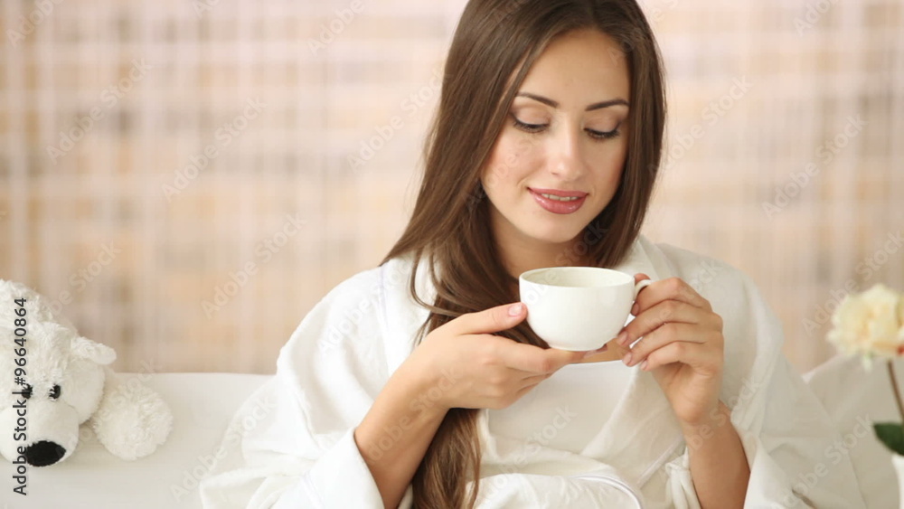 Attractive young woman relaxing in bed drinking tea looking at camera and smiling. Panning camera Attractive young woman relaxing in bed drinking tea looking at camera and smiling. Panning camera