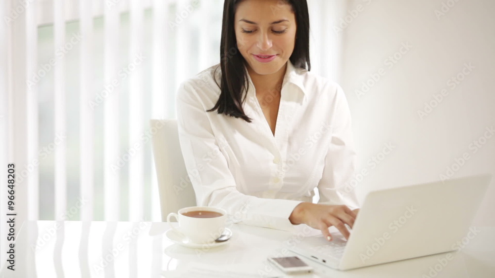 Cheerful young woman sitting at office desk using laptop stirring tea looking at camera and smiling. Panning camera Cheerful young woman sitting at office desk using laptop stirring tea looking at camera and smiling. Panning camera