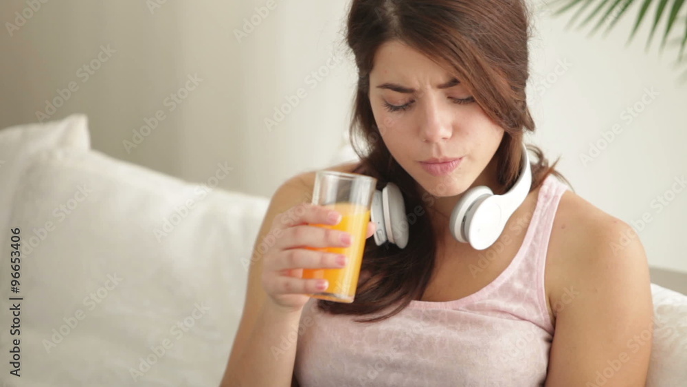 Charming girl wearing headset sitting on sofa using mobile phone drinking juice looking at camera and smiling. Panning camera Charming girl wearing headset sitting on sofa using mobile phone drinking juice looking at camera and smiling. Panning camera