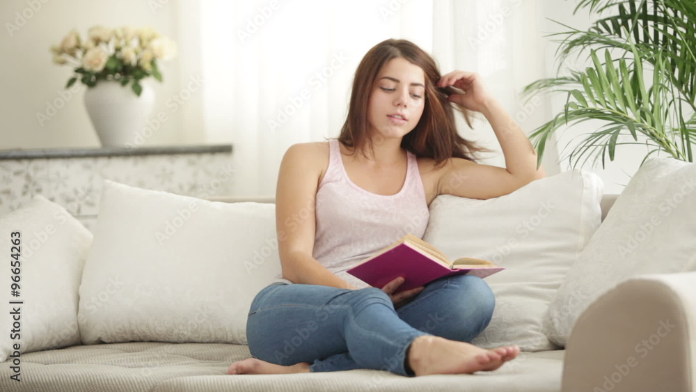 Cute girl sitting on sofa reading book looking at camera and smiling Cute girl sitting on sofa reading book looking at camera and smiling