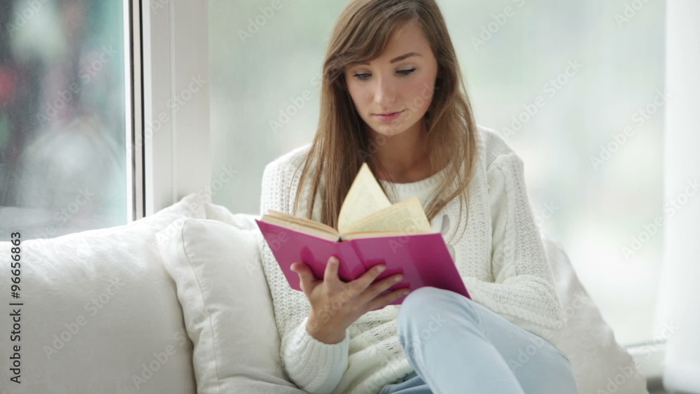 Cute girl sitting by window reading book looking at camera and smiling. Panning camera Cute girl sitting by window reading book looking at camera and smiling. Panning camera